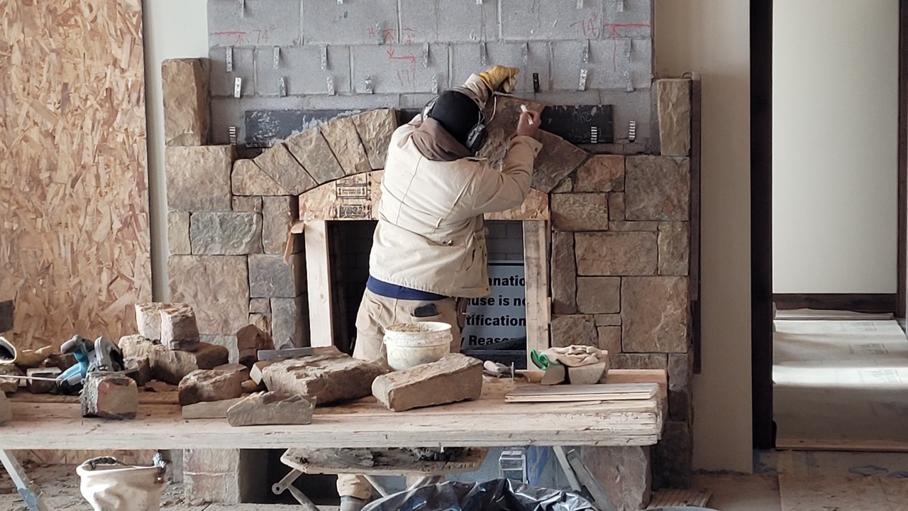 A construction worker in a jacket and gloves is building a stone fireplace in an unfinished room, using a trowel to apply mortar.